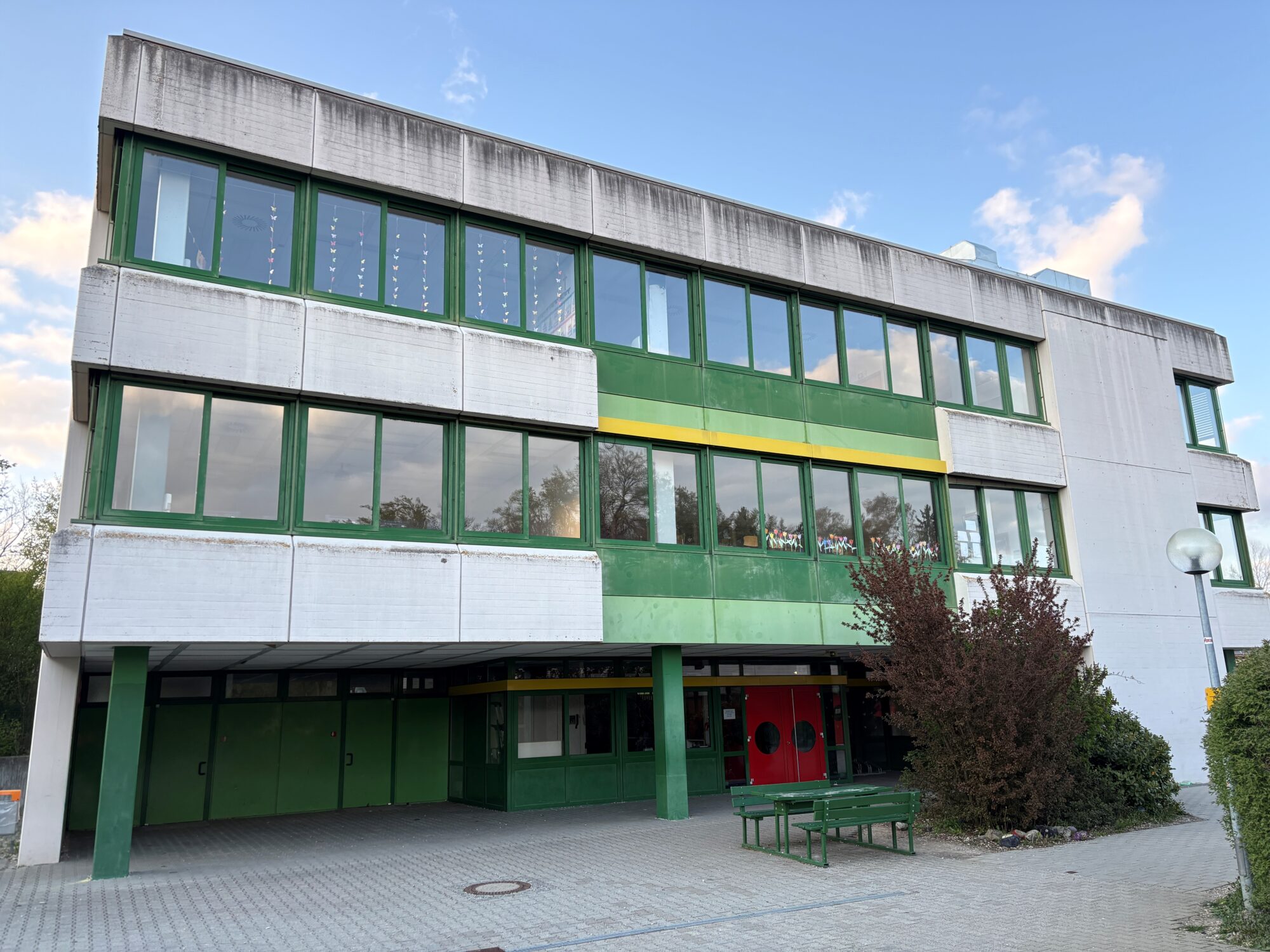 Ein dreistöckiges Gebäude mit weißen und grünen Fassadenplatten, großen Fenstern und einem Flachdach. Davor stehen eine Bank und einige Sträucher, über ihnen erstreckt sich ein größtenteils klarer, teilweise bewölkter Himmel.