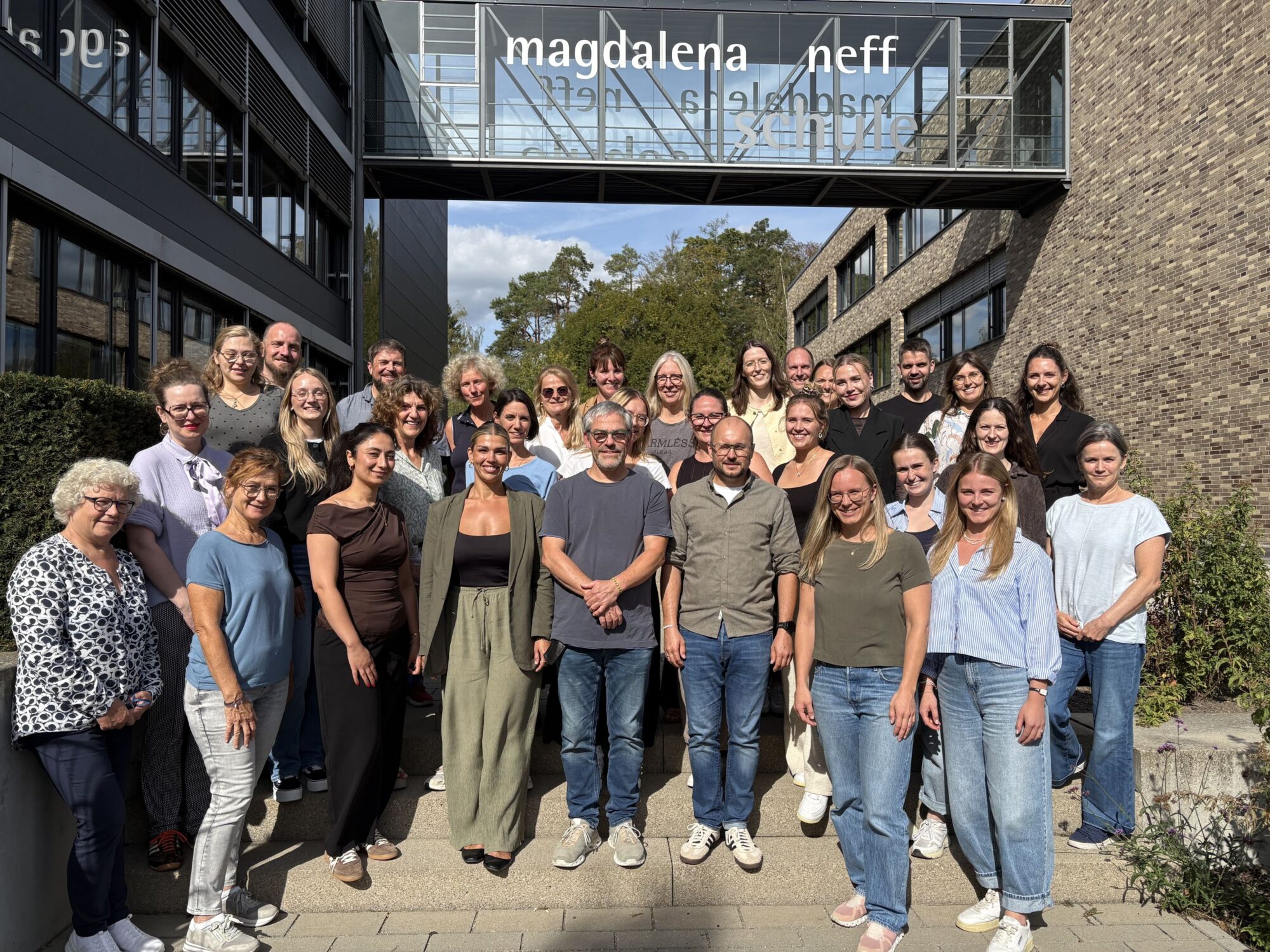 Eine Gruppe von etwa 30 Personen, die lächelnd draußen zwischen modernen Gebäuden stehen, über ihnen ein Schild mit der Aufschrift „magdalena neff“. Es ist sonnig, und alle sind leger gekleidet.