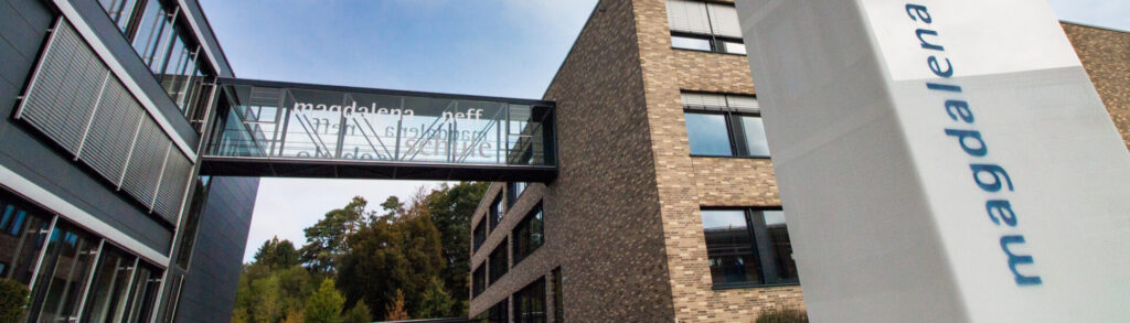 Ein modernes Gebäude mit einer Skybridge, die zwei Bereiche miteinander verbindet. Der Schriftzug "magdalena" ist auf einer weißen vertikalen Struktur im Vordergrund und auf der gläsernen Skybridge gut sichtbar angebracht. Im Hintergrund sind Bäume zu sehen.
