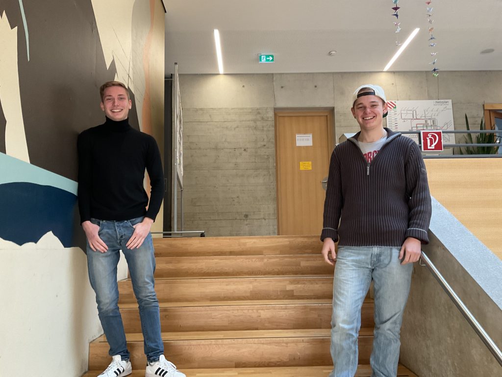 Zwei junge Männer stehen auf einer Holztreppe in einem Innenraum. Der eine links trägt einen schwarzen Rollkragenpullover und Jeans, der andere rechts einen braunen Pullover, Jeans und eine Mütze. Sie lächeln.