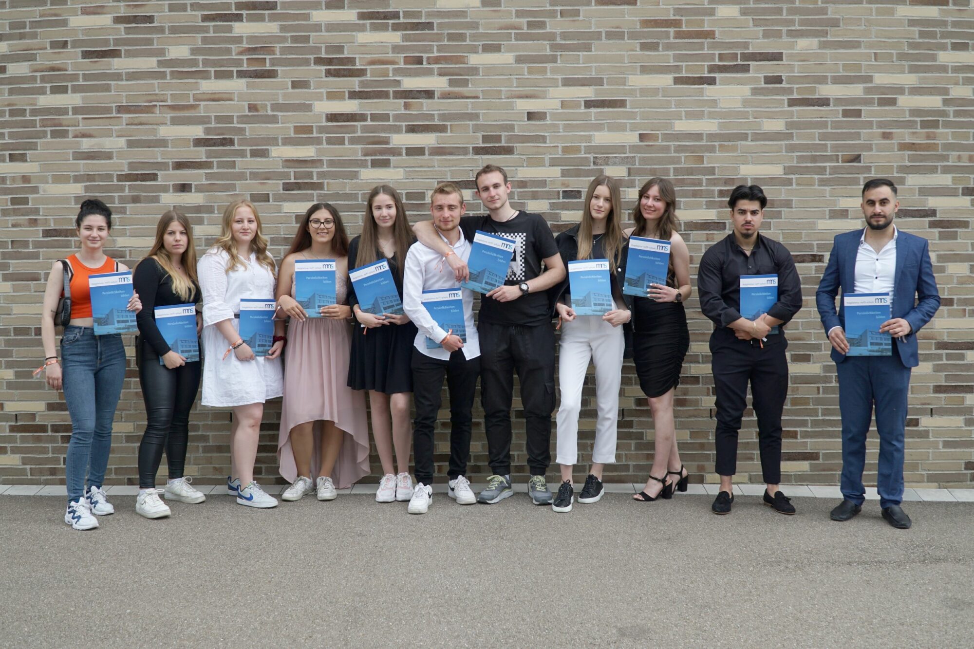 Eine Gruppe von elf jungen Erwachsenen steht in einer Reihe vor einer Backsteinmauer, jeder hält eine blaue Urkunde und posiert für ein Foto. Sie tragen eine Mischung aus legerer und halboffizieller Kleidung.