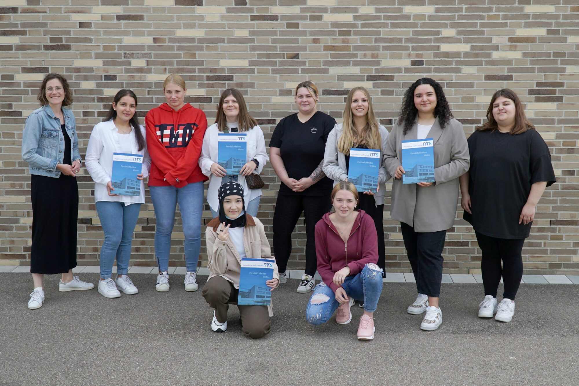 Eine Gruppe von elf Frauen posiert vor einer Backsteinmauer, einige stehen, andere knien. Viele halten Urkunden in der Hand und lächeln in die Kamera. Sie sind leger mit Jacken, Kapuzenpullis und Turnschuhen gekleidet.