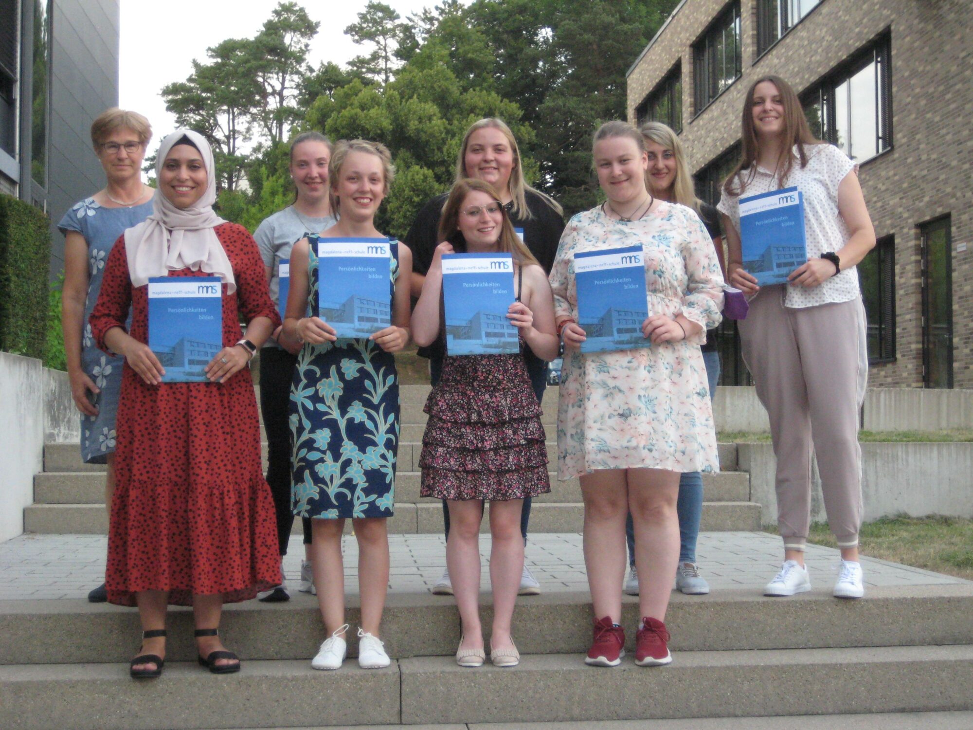 Eine Gruppe von acht jungen Frauen und einer älteren Frau steht im Freien auf einer Treppe, hält Urkunden in der Hand und lächelt, während im Hintergrund Bäume und moderne Gebäude zu sehen sind.