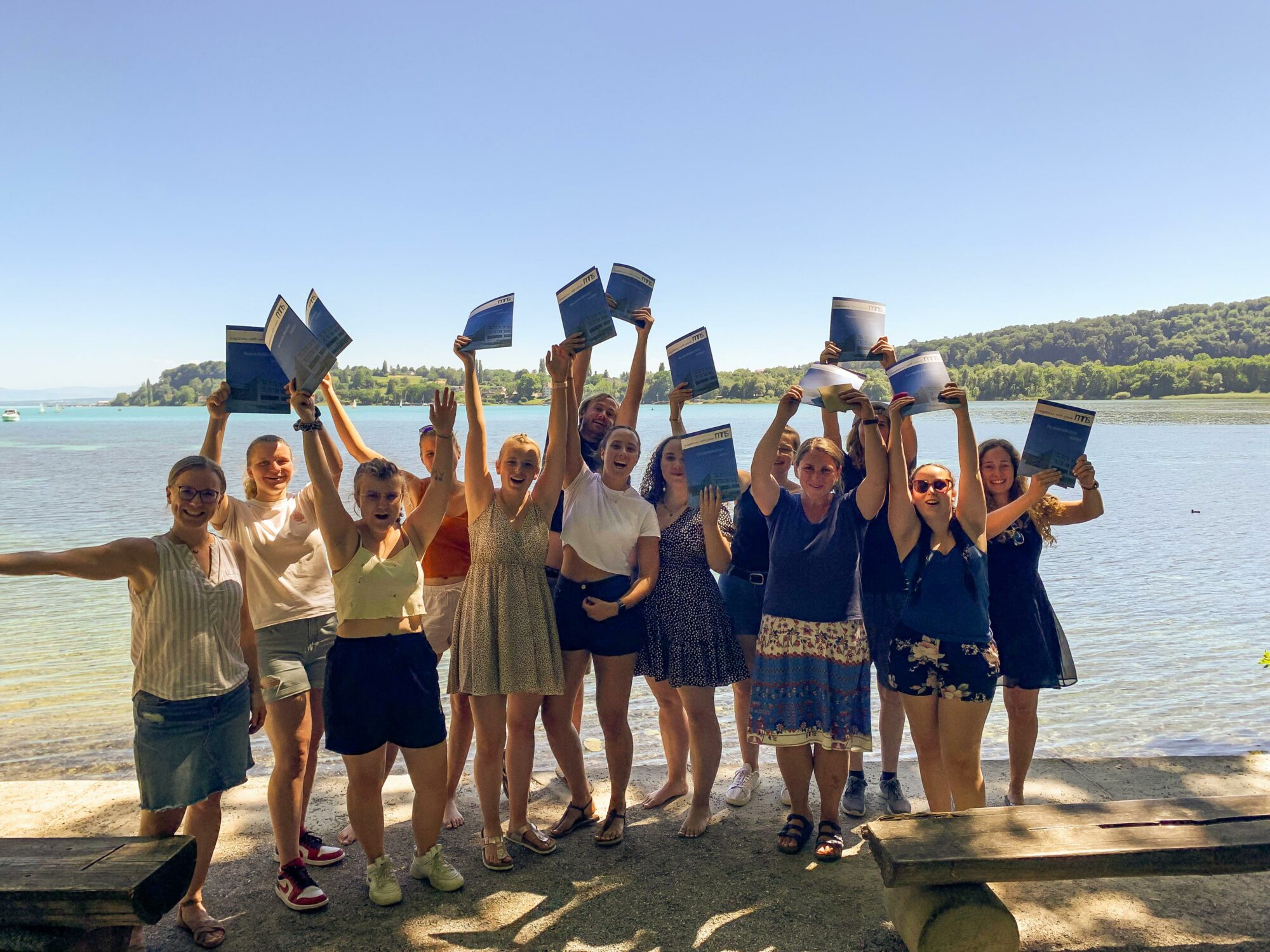 Eine Gruppe von Menschen steht an einem See, hält fröhlich lächelnd Urkunden oder Dokumente über ihren Köpfen, während im Hintergrund Bäume und klares blaues Wasser zu sehen sind.
