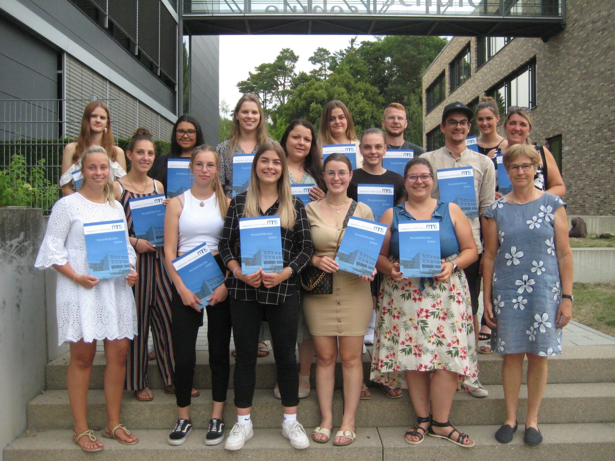 Eine Gruppe junger Erwachsener und zwei ältere Frauen stehen auf einer Freitreppe, lächeln und halten Urkunden in der Hand. Im Hintergrund sind moderne Gebäude und Grünflächen zu sehen. Alle sind leger gekleidet, und es scheint eine Abschluss- oder Preisverleihungsfeier zu sein.