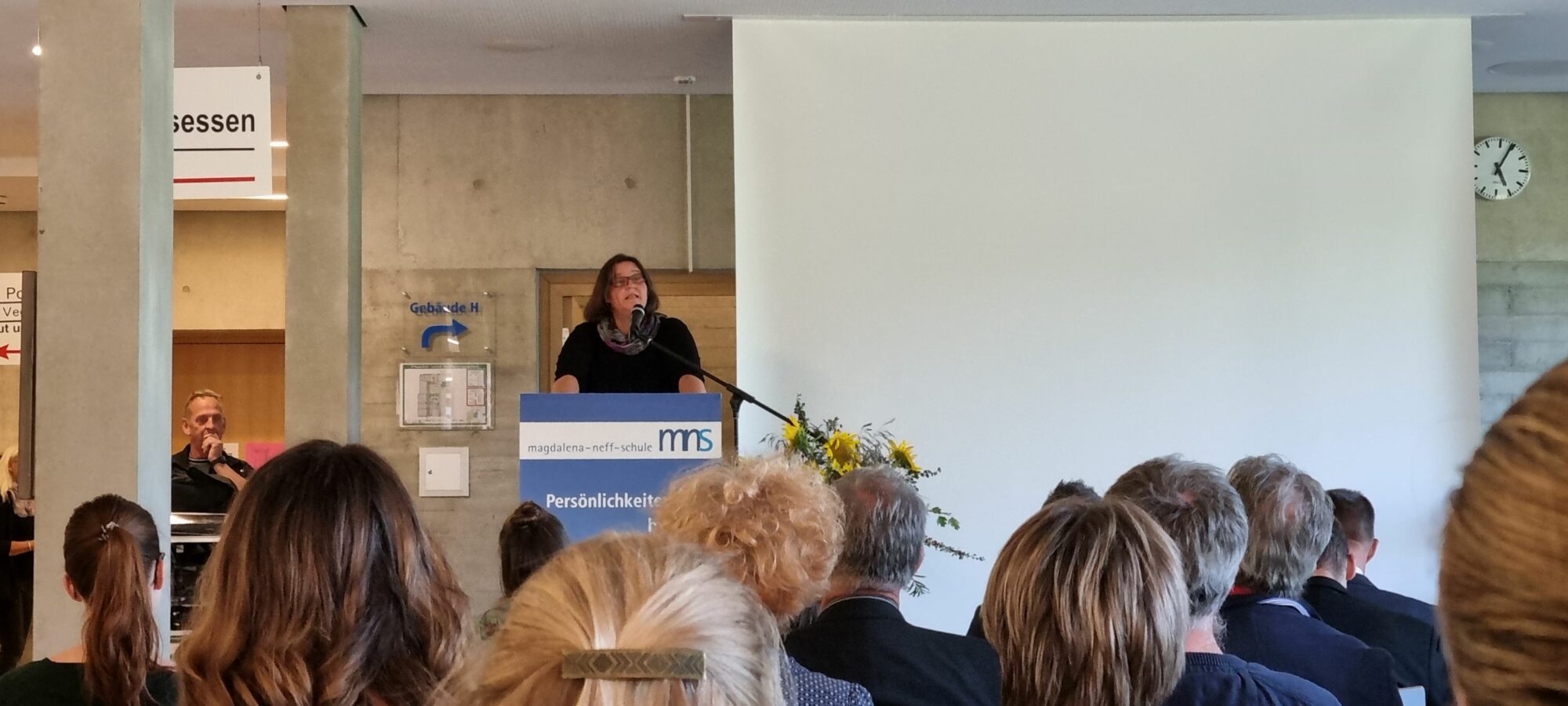 Eine Frau steht an einem Podium und spricht zu einem Publikum in einem Konferenzraum; hinter ihr sind eine Projektionsfläche und eine Uhr zu sehen, während im Vordergrund Menschen sitzen und zuhören.