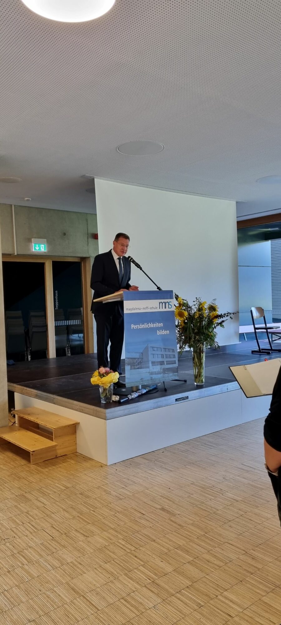 Ein Mann im Anzug steht an einem Podium auf einer kleinen Bühne und spricht zu einem Publikum. Neben dem Podium und auf dem Boden liegen Blumen. Im Hintergrund sind eine weiße Leinwand und leere Stühle zu sehen.