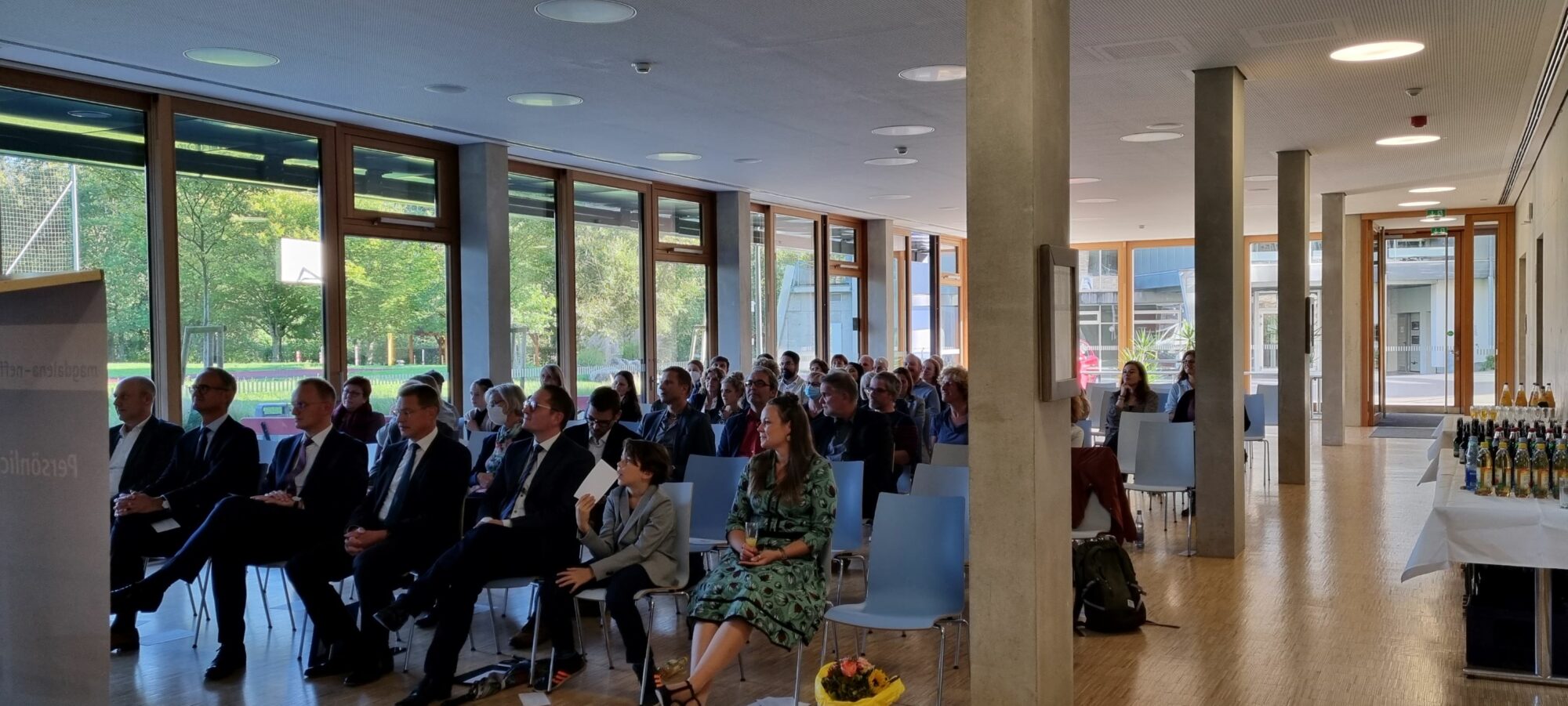 Ein formell gekleidetes Publikum sitzt in Stuhlreihen in einem hellen, modernen Raum mit großen Fenstern und hört einem Vortrag zu. Auf einem Tisch auf der rechten Seite stehen Getränke in Flaschen bereit.