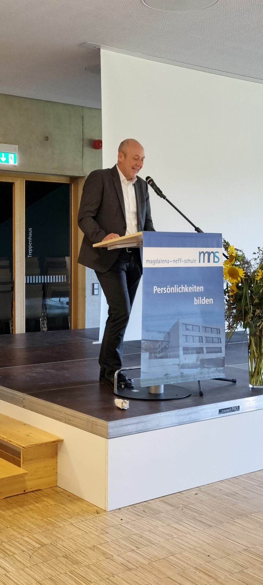 Ein Mann im Anzug steht an einem Podium auf einer Bühne und spricht in ein Mikrofon. Auf dem Podium stehen die Worte magdalena-neff-schule und Persönlichkeiten bilden. Ein Blumenstrauß steht neben dem Podium.