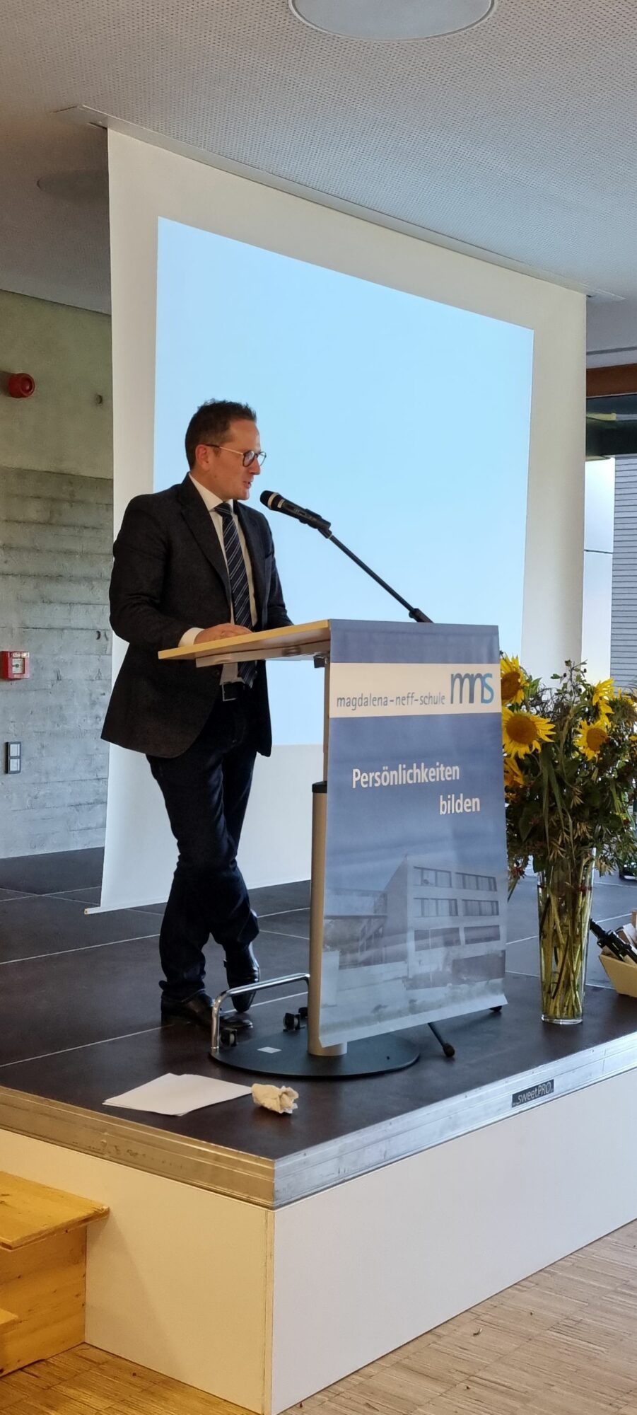 Ein Mann im Anzug spricht an einem Podium auf einer Bühne, hinter ihm ein Bildschirm, daneben ein großer Sonnenblumenstrauß. Auf dem Podium steht ein Schild mit der Aufschrift magdalena-neff-schule und Persönlichkeiten bilden.