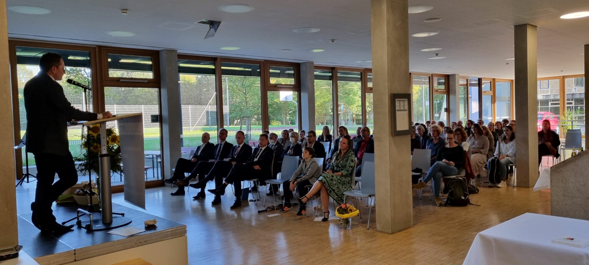 Ein Mann spricht von einem Podium aus zu einem sitzenden Publikum in einem hellen, modernen Raum mit großen Fenstern und Blick ins Grüne. Die Anwesenden, die sowohl geschäftlich als auch leger gekleidet sind, hören aufmerksam zu.
