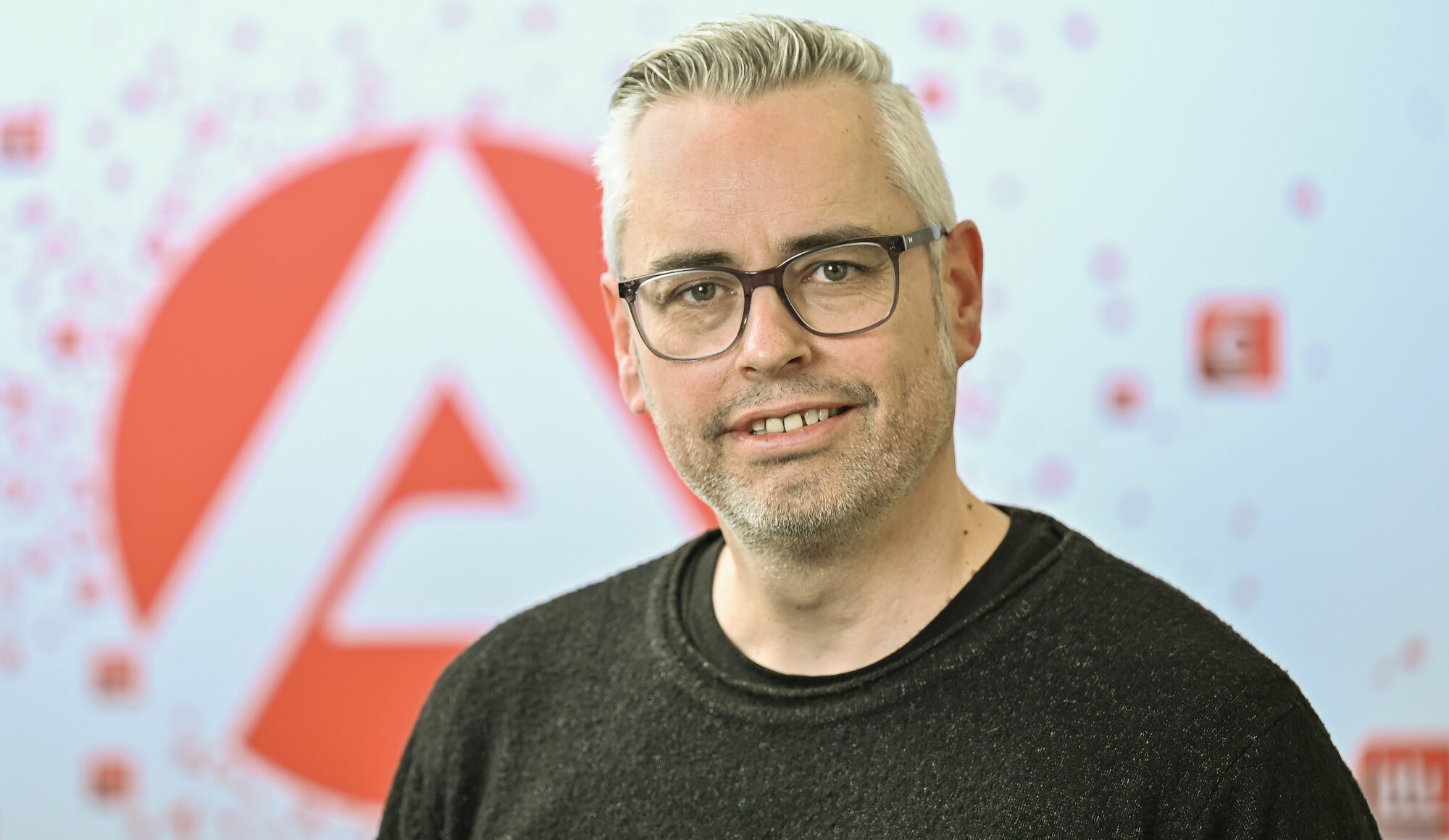 Ein Mann mit kurzen grauen Haaren, Brille und einem dunklen Pullover lächelt in die Kamera. Im Hintergrund ist ein großes rotes und weißes geometrisches Muster mit verstreuten Symbolen zu sehen.