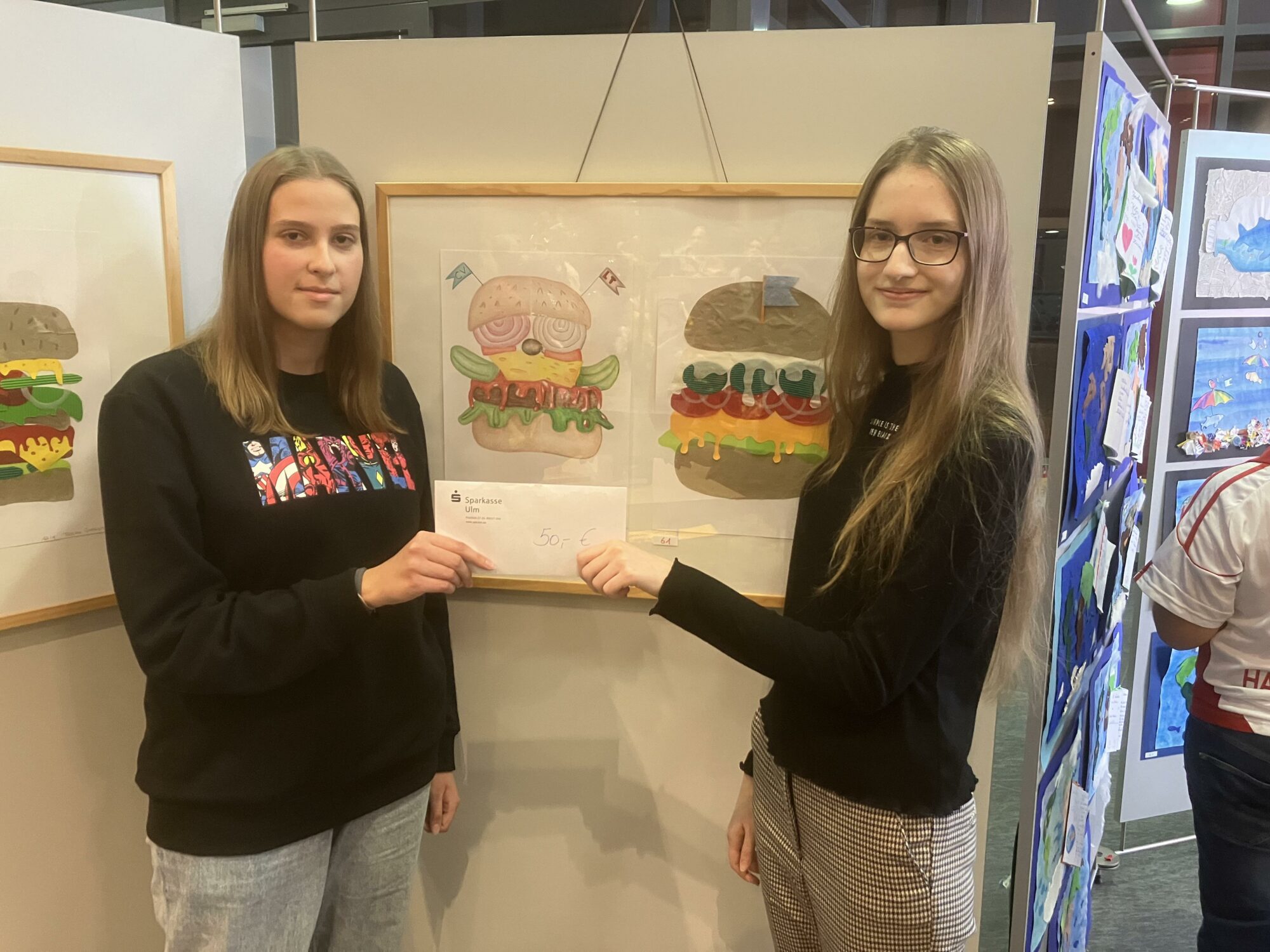 Zwei junge Frauen stehen neben einer gerahmten Zeichnung von Cartoon-Burgern und halten gemeinsam ein Zertifikat oder einen Umschlag in einer Kunstausstellung mit verschiedenen bunten Kunstwerken im Hintergrund.