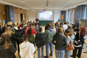 Eine Gruppe von Personen steht in einem Klassenzimmer und blickt auf einen Vortragenden, der vor einer großen Leinwand mit einer projizierten Folie spricht. Eine weitere Person steht in der Nähe, und an einer Wand sind Plakate zu sehen.
