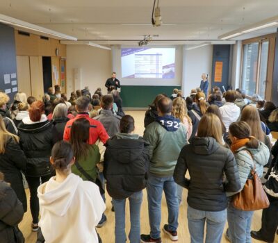Eine Gruppe von Personen steht in einem Klassenzimmer und blickt auf einen Vortragenden, der vor einer großen Leinwand mit einer projizierten Folie spricht. Eine weitere Person steht in der Nähe, und an einer Wand sind Plakate zu sehen.