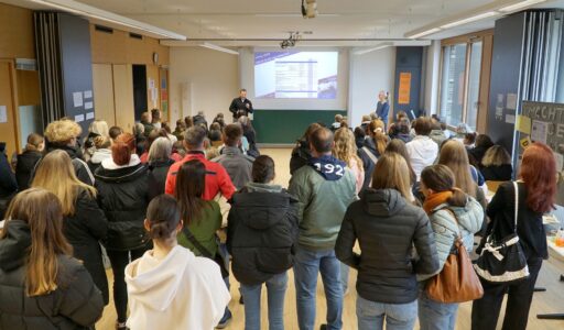Eine Gruppe von Personen steht in einem Klassenzimmer und blickt auf einen Vortragenden, der vor einer großen Leinwand mit einer projizierten Folie spricht. Eine weitere Person steht in der Nähe, und an einer Wand sind Plakate zu sehen.